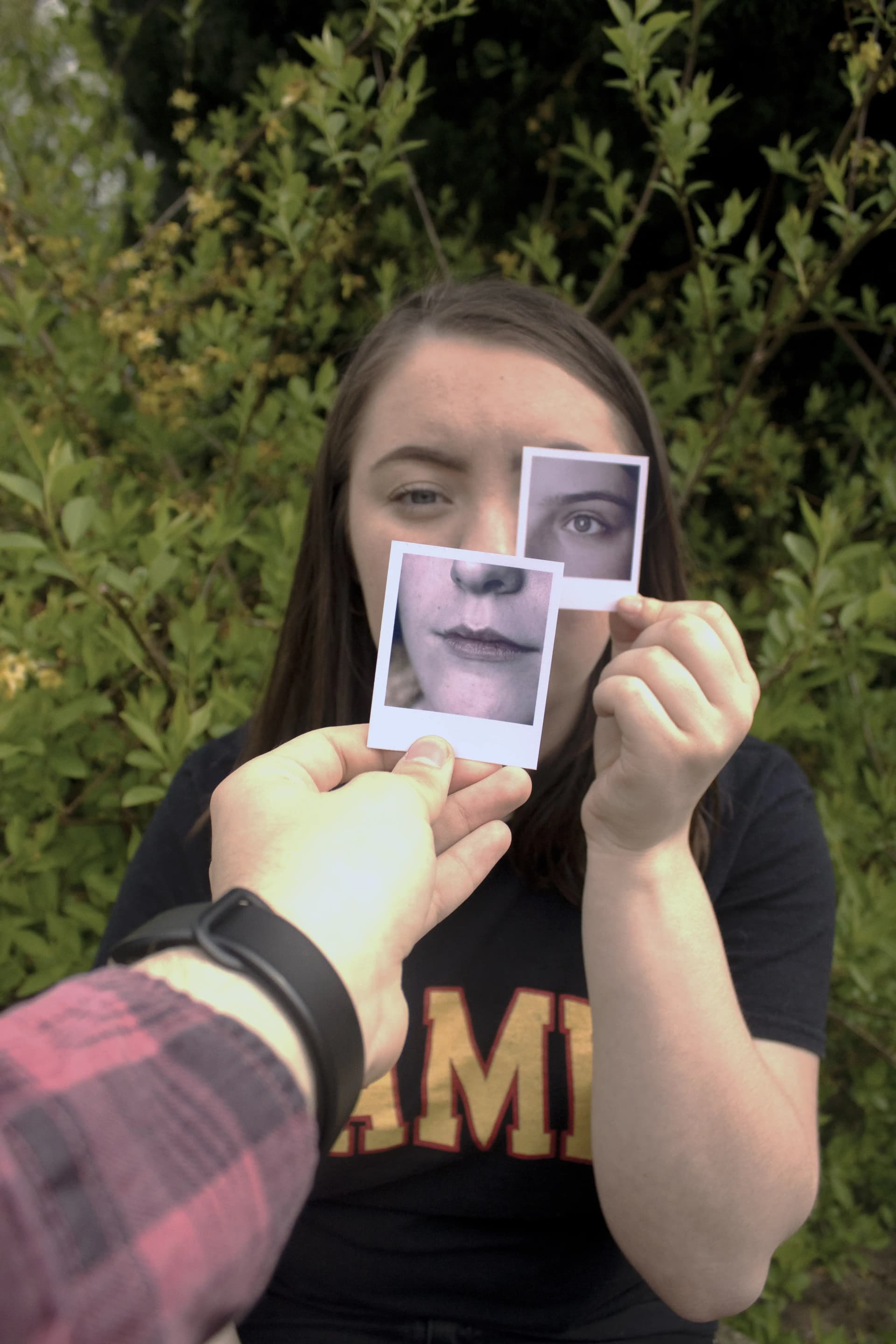 A person holds up two small Polaroid photographs in front of their face, aligning them with their own features. One photo shows their mouth and chin, held over their actual mouth and chin. The other photo shows their eye, positioned over their right eye.
