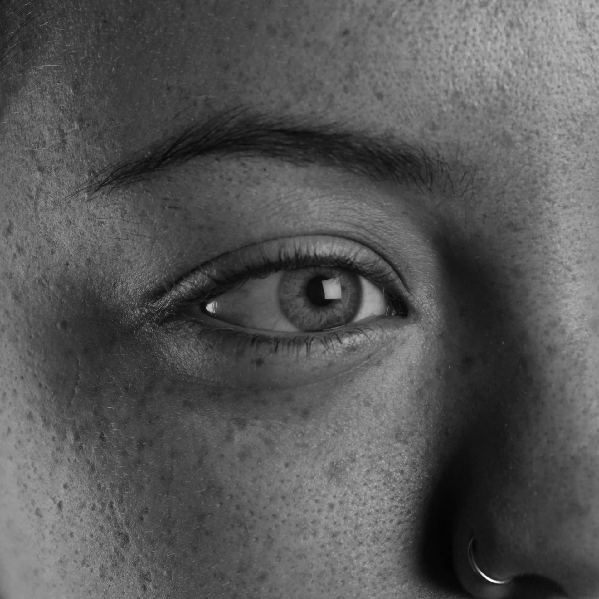 A detailed black and white close-up photograph showcases a human eye, slightly angled to the right. The intricate patterns of the iris are clearly visible, surrounding a dark pupil with a bright reflection. Delicate eyelashes extend from the lower eyelid, and the upper lid casts a subtle shadow. A defined eyebrow arches above the eye. The surrounding skin reveals texture and several small freckles, and a portion of a nose with a ring piercing is visible in the lower right corner.