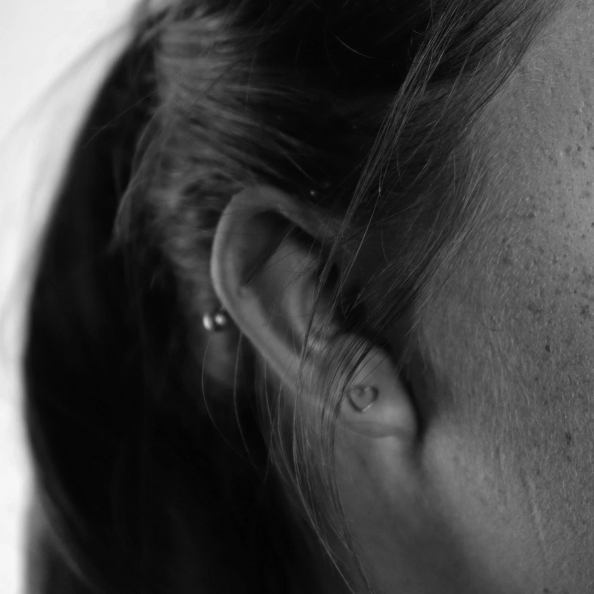 A detailed black and white close-up photograph showcases a human ear in profile. The intricate curves and folds of the outer ear, including the helix and lobule, are clearly defined. Two small, round earrings are visible, one in the lobe and another higher up on the helix. Wisps of dark hair frame the ear, and the texture of the skin on the side of the face is also visible, showing subtle variations in tone and a few small blemishes.