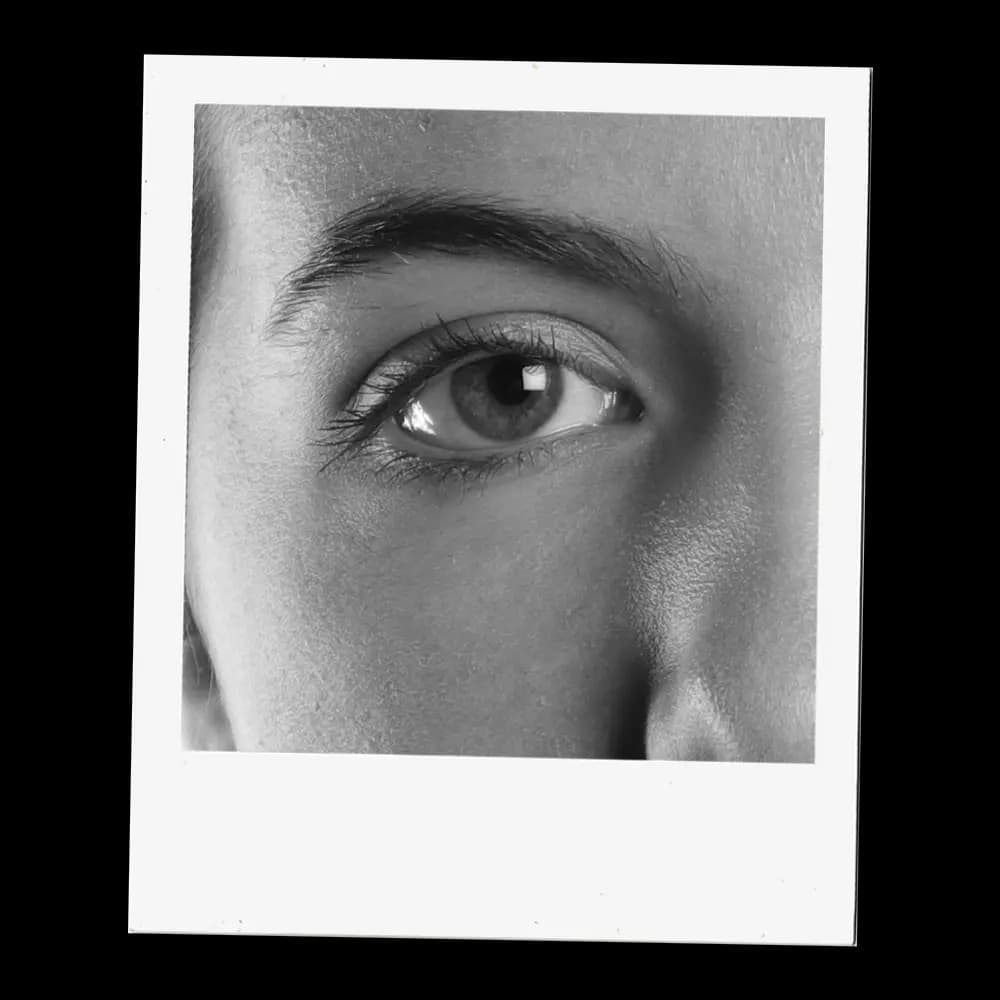 A close-up, black and white photograph captures the intricate details of a human eye. The iris, a mesmerizing mix of light and dark tones, surrounds a dark pupil, reflecting subtle highlights. Delicate eyelashes frame the lower eyelid, while the upper lid is partially visible, casting a soft shadow. Above the eye, a well-defined eyebrow adds structure to the face. The surrounding skin shows fine texture and subtle shading, emphasizing the eye as the focal point.