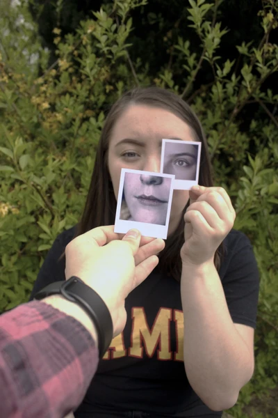 A person holds up two small Polaroid photographs in front of their face, aligning them with their own features. One photo shows their mouth and chin, held over their actual mouth and chin. The other photo shows their eye, positioned over their right eye. A person holds up two small Polaroid photographs in front of their face, aligning them with their own features. One photo shows their mouth and chin, held over their actual mouth and chin. The other photo shows their eye, positioned over their right eye.