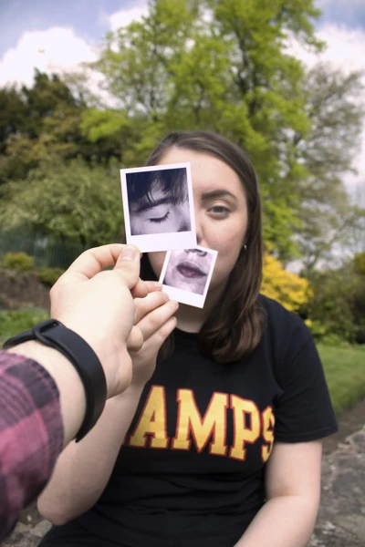 A person holds up two small Polaroid photographs in front of their face, aligning them with their own features. One photo shows their mouth and chin, held over their actual mouth and chin. The other photo shows their eye, positioned over their left eye. A person holds up two small Polaroid photographs in front of their face, aligning them with their own features. One photo shows their mouth and chin, held over their actual mouth and chin. The other photo shows their eye, positioned over their left eye.