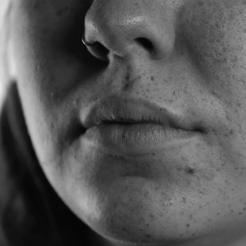A detailed black and white close-up photograph focuses on the lower portion of a human face. The subject's full lips are centrally positioned, showing their texture and subtle contours. Above the lips, the base of a nose is visible, featuring a ring piercing in the left nostril. The surrounding skin exhibits a slightly uneven texture and a few small blemishes. The overall lighting highlights the subtle variations in tone and surface detail. A detailed black and white close-up photograph focuses on the lower portion of a human face. The subject's full lips are centrally positioned, showing their texture and subtle contours. Above the lips, the base of a nose is visible, featuring a ring piercing in the left nostril. The surrounding skin exhibits a slightly uneven texture and a few small blemishes. The overall lighting highlights the subtle variations in tone and surface detail.